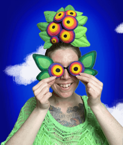 Portrait of artist wearing green, head and eyes covered in colorful eye-like creations, animated clouds looping in background.